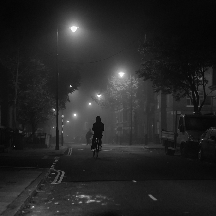 A hooded person is riding a bike in London at night in the road.
