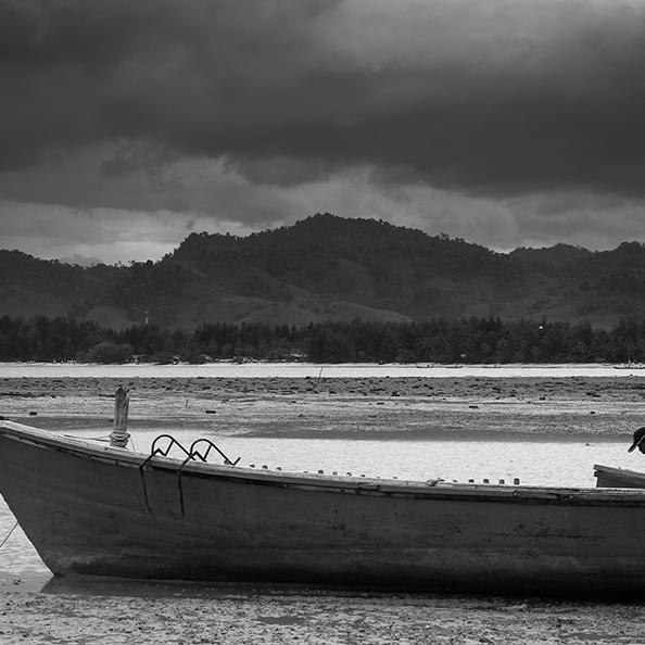 A black and white photo of a man sat in boat on beach in Thialnad with hills in the background.